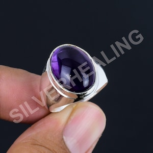 Könnte beinhalten: Ein Silberring mit einem großen, ovalen, tiefvioletten Amethyst-Edelstein, der in ein glattes, poliertes Metallband gefasst ist. Das Wort SILVERHEALING ist diagonal in einer hellgrauen, halbtransparenten Schrift über das Bild gedruckt.