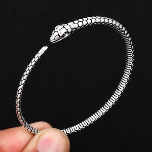 Puede incluir: Pulsera de serpiente de plata con un diseño de serpiente texturizado. La pulsera está abierta y tiene una cabeza de serpiente en un extremo.