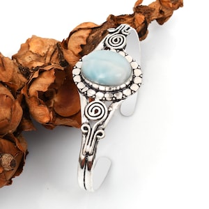 Può includere: Un bracciale a polsino argentato con una pietra preziosa ovale azzurra. La pietra è incastonata in una cornice dettagliata e bordata di perline. Il bracciale ha un design aperto con decorazioni a spirale e volute.