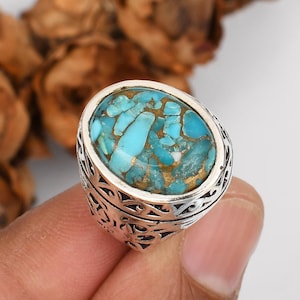 May include: A silver ring with an oval turquoise stone. The stone is a mosaic of light blue and gold-colored fragments. The ring has an ornate, openwork design around the band. The ring is held by a person.