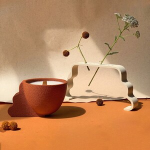 May include: A terracotta candle holder with a white candle and a wooden wick. The holder is shaped like a cloud and has a textured surface. The candle is sitting on a brown surface with a white ceramic flower vase with white flowers and brown stems.