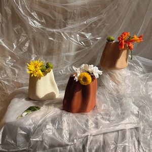 May include: Three ceramic vases with different textures and colours, each holding a bouquet of flowers. The vases are arranged on a white fabric surface with a plastic sheet draped over it.