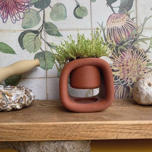 May include: A terracotta-coloured ceramic planter with a unique, rounded design. The planter has a small opening in the centre, where a smaller terracotta pot sits, holding a green plant.