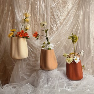 May include: Three ceramic vases with different coloured glazes, each holding a bouquet of flowers. The vases are sitting on a white fabric background.