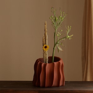 May include: A terracotta-colored ceramic vase with a textured, wavy design. The vase holds dried wheat, a small yellow flower, and a green plant with small buds. The arrangement sits on a dark wooden surface.