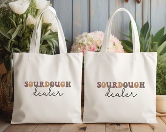 May include: Two white tote bags with the words "SOURDOUGH dealer" printed in brown, tan, and black. The bags have white handles and are sitting on a wooden surface. Flowers and plants are in the background.