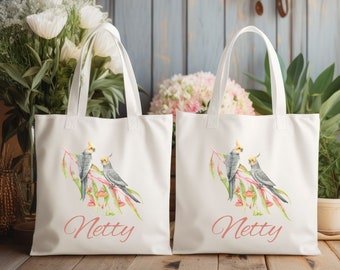 May include: Two white tote bags with a pink floral design and two grey cockatiels on each bag. The bags have the name "Netty" printed on them in pink.
