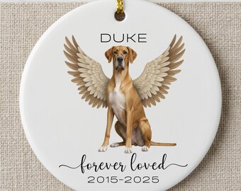 May include: A white ceramic ornament with a golden retriever illustration wearing angel wings. The ornament has the name "DUKE" above the dog and the text "forever loved" and dates "2015-2025" below the dog. A gold string is attached for hanging.