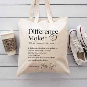 Peut inclure: Un sac fourre-tout en toile blanche avec du texte noir qui dit "Difference Maker" et une définition du mot. Le texte comprend également le nom "Mrs. Taylor".