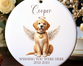May include: A white ceramic ornament with a golden retriever angel illustration. The dog has wings, a halo, and a heart-shaped pendant. The name "Cooper" is written above the dog, and the text "WISHING YOU WERE HERE 2012-2024" is below.