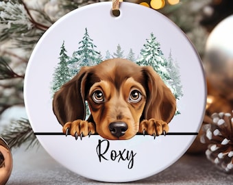 May include: A round, white ceramic Christmas ornament featuring a brown and tan dachshund dog with big eyes, paws resting on a black line. The dog is in front of a snowy forest scene. The name "Roxy" is written in cursive.