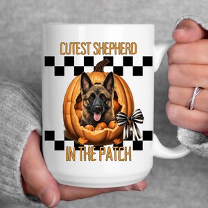 Peut inclure: Mug en céramique blanche avec un berger allemand à l'intérieur d'une citrouille sculptée. Le mug porte l'inscription "CUTEST SHEPHERD IN THE PATCH" en orange. Un motif à carreaux noir et blanc encadre le motif de la citrouille.