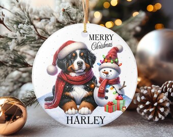 May include: Round Christmas ornament featuring a Bernese Mountain Dog puppy and a snowman, both wearing Santa hats and red scarves. The ornament has the text "Merry Christmas" and "Harley".