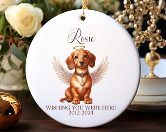 May include: A white ceramic ornament with a gold hanging cord. The ornament features a brown dachshund with angel wings and a halo. The text "Rosie" is above the dog, and "WISHING YOU WERE HERE 2012-2024" is below.
