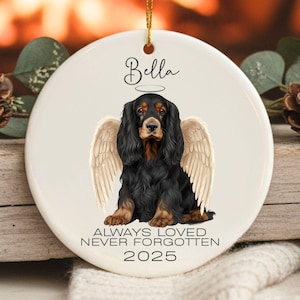 May include: A white ceramic ornament featuring a black and brown dog with angel wings and a halo. The dog's name, "Bella," is above it, and the text "ALWAYS LOVED NEVER FORGOTTEN 2025" is below. A gold string is attached for hanging.