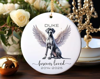 May include: A white ceramic ornament featuring a Great Dane with angel wings, the name "DUKE" above, and "forever loved 2014-2025" below. The ornament hangs from a gold chain. The background includes flowers and gold accents.