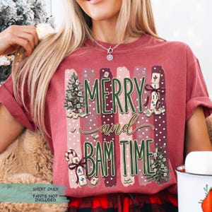May include: A brick red t-shirt featuring a graphic print with the text MERRY and BAM TIME. The design includes vertical stripes, snowflakes, two Christmas trees decorated with mahjong tiles, and stacks of mahjong tiles tied with ribbons.