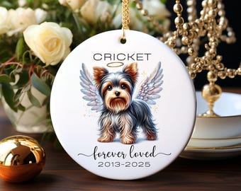 May include: A circular white ceramic ornament with a gold chain. It features a Yorkshire Terrier with angel wings, the name "Cricket", and the text "forever loved 2013-2025". The ornament is designed to be hung and is a memorial item.