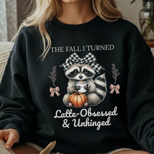 May include: Black sweatshirt featuring a raccoon with a bow, holding a coffee cup and sitting on a pumpkin. Text reads "The Fall I Turned Latte-Obsessed & Unhinged". The design includes fall-themed accents.