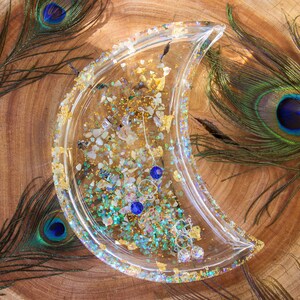 Può includere: Un vassoio di resina trasparente a forma di mezzaluna riempito di glitter iridescenti, foglie d'oro e piccole pietre preziose. Il vassoio contiene diversi paia di orecchini con accenti blu e cristallo trasparente. Il vassoio è appoggiato su una superficie di legno, con piume di pavone sullo sfondo.