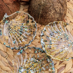 Op de afbeelding: Drie doorzichtige schelpvormige schalen van hars met gouden en iriserende glitter en vlokken. De schalen liggen op een geweven mat met twee bruine kokosnoten op de achtergrond. De schalen zijn ongeveer 10 cm breed.