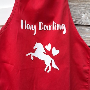 May include: Red apron with white text that reads "Hay Darling". A white silhouette of a horse and two hearts are below the text. The apron has a pocket and neck and waist ties.