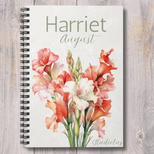 May include: A spiral-bound notebook with a floral design. The cover features the name "Harriet" and the month "August" in green script. The floral arrangement includes red, orange, and white gladiolus flowers. The word "Gladiolus" is written at the bottom.