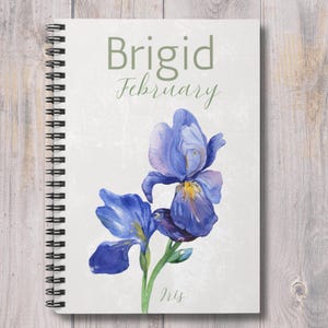 May include: A spiral bound notebook with a white cover featuring a watercolor painting of a purple iris flower. The cover also includes the text "Brigid February Iris".