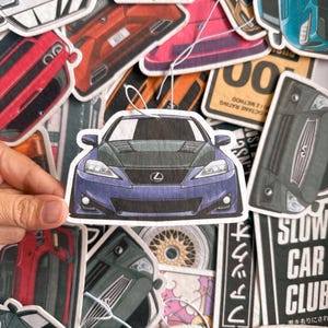 以下が含まれることがあります： 車のテーマの芳香剤のコレクション。主なものは、黒いボンネットが付いた青いレクサスの車の芳香剤です。他の芳香剤は、さまざまな車のデザインと「SLOW CAR CLUB」のテキストを表示しています。