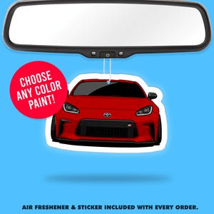 Peut inclure: Un désodorisant pour voiture rouge en forme de voiture, suspendu à un rétroviseur. La voiture a des détails noirs et un emblème Toyota. Un cercle rouge avec le texte "CHOOSE ANY COLOR PAINT!" est à gauche. Le fond est bleu.