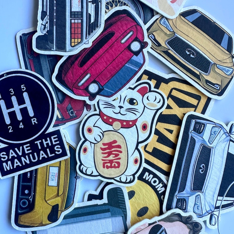以下が含まれることがあります： 車のイラスト、招き猫、テキストが特徴のカラフルなステッカーのコレクションです。「SAVE THE MANUALS」と書かれたステッカー、赤い車、金色の車が含まれています。