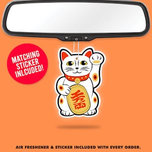 May include: White ceramic car air freshener shaped like a Japanese Maneki-Neko, or lucky cat, with a raised paw and a gold coin with the Chinese character for "fortune" on its chest. The air freshener is hanging from a rearview mirror. A red circle with the text "MATCHING STICKER INLCUDED!" is in the lower left corner of the image.