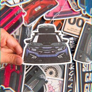 以下が含まれることがあります： 青と黒の車のイラストが描かれた、車のテーマの芳香剤。ルーフキャリア付き。芳香剤は手で持たれており、他の車関連のデザインに囲まれています。