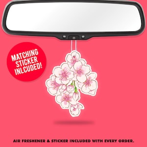 Könnte beinhalten: Ein Auto-Lufterfrischer in Form eines Clusters aus rosa und weißen Kirschblüten, der an einem Rückspiegel hängt. Ein roter Kreis mit dem Text "MATCHING STICKER INLCUDED!" ist sichtbar. Der Hintergrund ist einfarbig rosa.