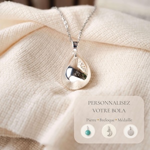 Peut inclure: Un collier avec un pendentif en forme de goutte argentée sur une chaîne fine. Un encart affiche le texte PERSONNALISEZ VOTRE BOLA, Pierre, Breloque, Médaille, avec des icônes montrant une perle turquoise, une breloque lune et un disque.