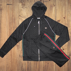 May include: A black zip-up hooded sweatshirt and matching track pants set. The hoodie features white piping along the raglan sleeves, a gold-toned interlocking logo on the chest, and green and red side stripes. The pants include matching side stripes.