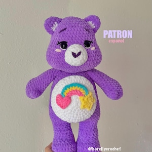 Puede incluir: Un peluche de oso tejido a crochet en color morado con un hocico blanco y un parche circular blanco en el vientre que muestra un arcoíris colorido, un corazón rosa y una estrella amarilla. El texto PATRON español y @karellycrochet son visibles.