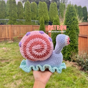 May include: A crocheted snail toy in shades of blue, pink, and purple. The snail's shell is a spiral of pink and brown, with purple dots. The snail's body is light blue, with a ruffled, mint green base. The text "PATRON @karellycrochet" is visible.