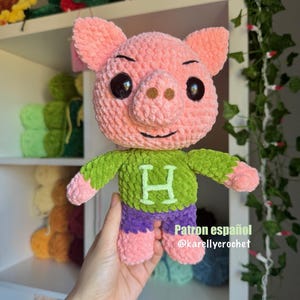 May include: A pink crocheted pig wearing a green sweater with a large green letter H on the front and purple trousers. The pig is being held by a hand. The text "Patron español @karellycrochet" is visible in the bottom right corner of the image.