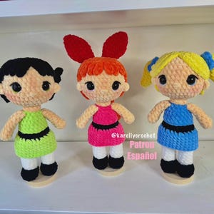 Puede incluir: Tres muñecas de ganchillo hechas a mano, cada una con un peinado y atuendo únicos. Las muñecas visten vestidos verdes, rosas y azules con detalles negros. Las muñecas están sobre bases de madera.