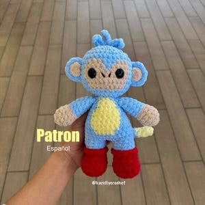 Puede incluir: Un mono de ganchillo azul hecho a mano con detalles beige en la cara, las manos y los pies. El mono tiene una barriga amarilla y botas rojas. El texto "Patron Español" es visible.