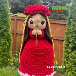May include: A crocheted doll featuring a red dress with a white scalloped hem, a red rose headpiece, and a green and gold shawl. The doll has brown hair, black eyes, and hands clasped in prayer holding a small silver cross. Text reads PATRON español.