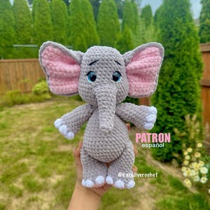 Puede incluir: Un peluche de elefante gris tejido a mano con orejas internas rosas y ojos azules. El elefante tiene detalles blancos en las patas y se sostiene frente a un fondo verde. El texto "PATRON español" y "@karellycrochet" son visibles.