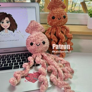 Puede incluir: Dos peluches de calamar tejidos a crochet, uno rosa y otro naranja, con ojos negros y bocas sonrientes. El calamar rosa está sentado en una computadora portátil con un gráfico de ovillo de lana y aguja en el trackpad. El texto "Patron @karellycrochet" es visible en la mesa.