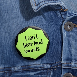 May include: A black and green button with the text "I can't bear loud sounds" on a denim jacket.