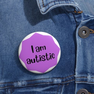 May include: A white button with a purple irregular shape in the center. The text "I am autistic" is printed in black. The button is pinned to a blue denim jacket.