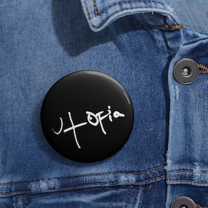 Puede incluir: Un botón negro y redondo con la palabra "Utopia" escrita en blanco. El botón brillante se encuentra sobre una chaqueta vaquera.