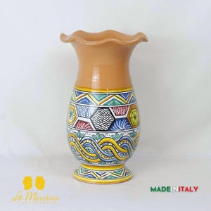 Puede incluir: Un jarrón de terracota con un borde festoneado y un patrón geométrico y ondulado colorido. El jarrón está decorado con pintura azul, verde, amarilla, roja y blanca.