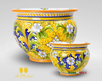 May include: Two ceramic flower pots with a vibrant yellow background and intricate floral designs in blue, white, and green. The pots have a rounded shape and are adorned with decorative patterns. The rim and base are orange. The text "MADE IN ITALY" is visible.