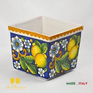 Può includere: Vaso quadrato in ceramica con sfondo blu, decorato con limoni gialli, fiori bianchi e foglie verdi. Il bordo è smerlato e dipinto in giallo dorato. Il vaso è etichettato "Made in Italy".